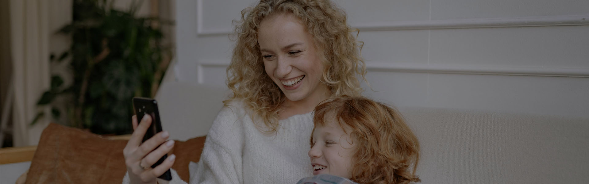 Mãe de camisa branca abraçada com a filha de camisa cinza deitadas no sofá olhando o celular que a mãe está segurando