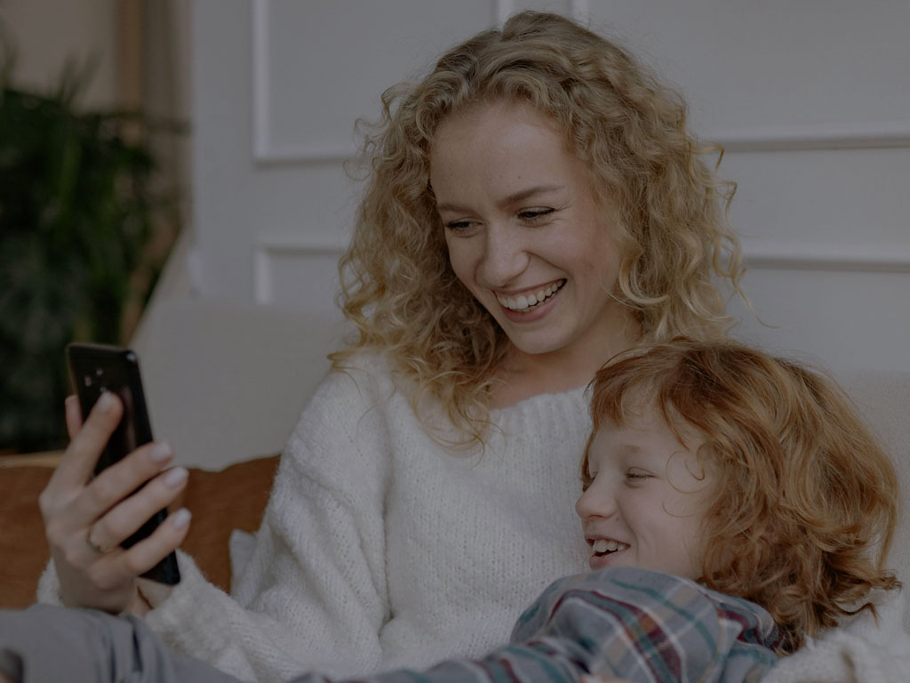 Mãe de camisa branca abraçada com a filha de camisa cinza deitadas no sofá olhando o celular que a mãe está segurando