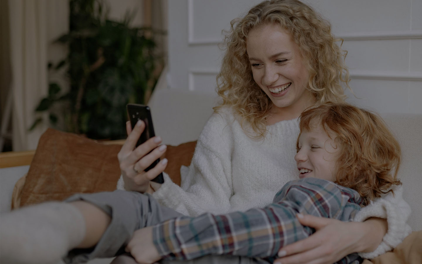 Mãe de camisa branca abraçada com a filha de camisa cinza deitadas no sofá olhando o celular que a mãe está segurando